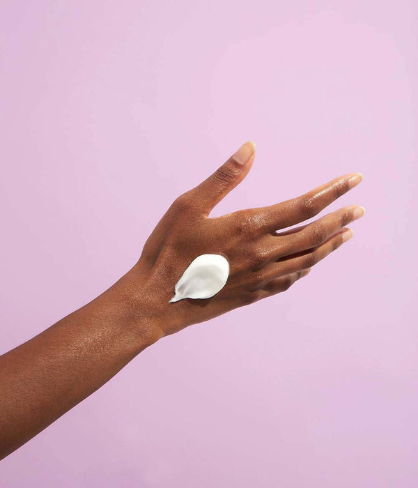 Deep skin tone model presenting a swatch of hand cream on the back of the hand against a purple background.